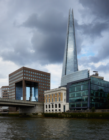 Dark shard This urban architecture photograph captures the London skyline in the late afternoon during the summer. The main subject of the image is The Shard, a prominent glass skyscraper in central London, which stands tall against dramatic summer clouds. In the foreground, the River Thames flows beside notable riverside buildings, including a historic structure and modern office complexes, with the characteristic city architecture well represented. The photograph provides a clear view of The Shard as a landmark, illustrating its imposing presence along the Thames and highlighting the contrast between old and new architectural styles that define this part of London.